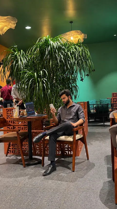 Meet Ranpara sitting in a caf&eacute;, reading on his phone beside a large plant and warm pendant lights