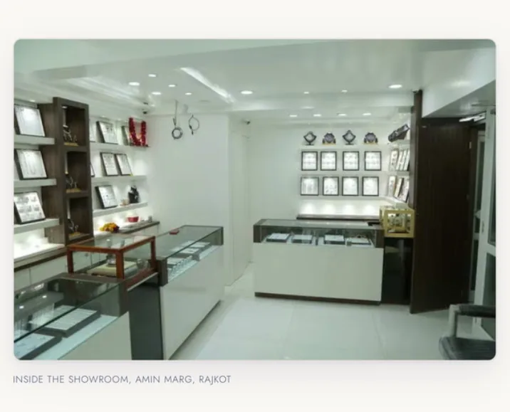 A photograph of the Drashti Jewels showroom interior on Amin Marg, Rajkot, with lit display counters and framed jewellery sets along the walls